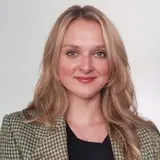 Porträt einer blonden Frau mit langen Haaren, die in einem karierten Blazer und einem schwarzen Shirt gekleidet ist. Sie lächelt freundlich in die Kamera. Der Hintergrund ist hell und neutral, was den Fokus auf ihr Gesicht lenkt.