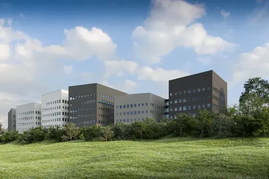 Mehrteiliges, modernes Gebäudekomplex mit rechteckigen Fassaden in unterschiedlichen Grautönen. Im Vordergrund eine grüne Wiese mit Büschen.