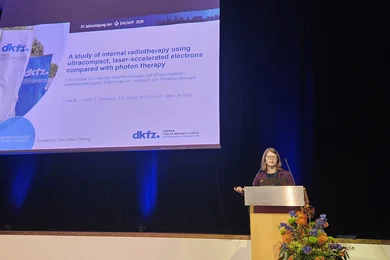 Eine Präsentatorin steht an einem Rednerpult auf einer Konferenz. Im Hintergrund ist ein Bildschirm mit einer wissenschaftlichen Folie zu sehen, die sich mit interner Radiotherapie mithilfe von ultrakompakten, laserbeschleunigten Elektronen im Vergleich zur Photonentherapie beschäftigt.