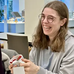Eine lächelnde Frau mit langen, lockigen Haaren und Brille hält eine Schüssel mit bunten Süßigkeiten in der Hand. Sie sitzt an einem Tisch in einem hellen Raum, umgeben von modernen Möbeln und Pflanzen.