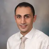 Ein Mann mit dunklen Haaren und braunen Augen trägt ein weißes Hemd und eine gestreifte Krawatte. Er lächelt freundlich in die Kamera vor einem neutralen Hintergrund.