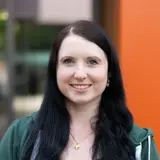 Eine Frau mit langen, schwarzen Haaren lächelt in die Kamera. Sie trägt ein schwarzes Oberteil mit Spitze und eine grüne Jacke. Im Hintergrund ist eine orangefarbene Wand zu sehen.