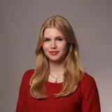 Eine blonde Frau mit langen Haaren lächelt in die Kamera. Sie trägt ein rotes Oberteil und eine Kette mit kleinen Anhängern. Im Hintergrund sind unscharfe Strukturen und Licht, die eine städtische Umgebung vermuten lassen.