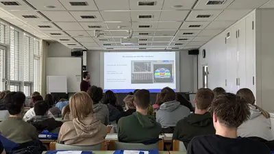 Ein Dozent steht vor einer Projektorleinwand in einem Klassenraum und erklärt ein Thema, während eine Gruppe von Schülern aufmerksam zuhört. Auf dem Bildschirm sind Grafiken und Informationen zu sehen, die für die Präsentation relevant sind.