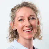 Ein Porträt einer Frau mit lockigem, blondem Haar, die in die Kamera lächelt. Sie trägt ein hellblaues Hemd und Ohrstecker. Der Hintergrund ist neutral und hell, wodurch ihr Gesicht gut zur Geltung kommt.