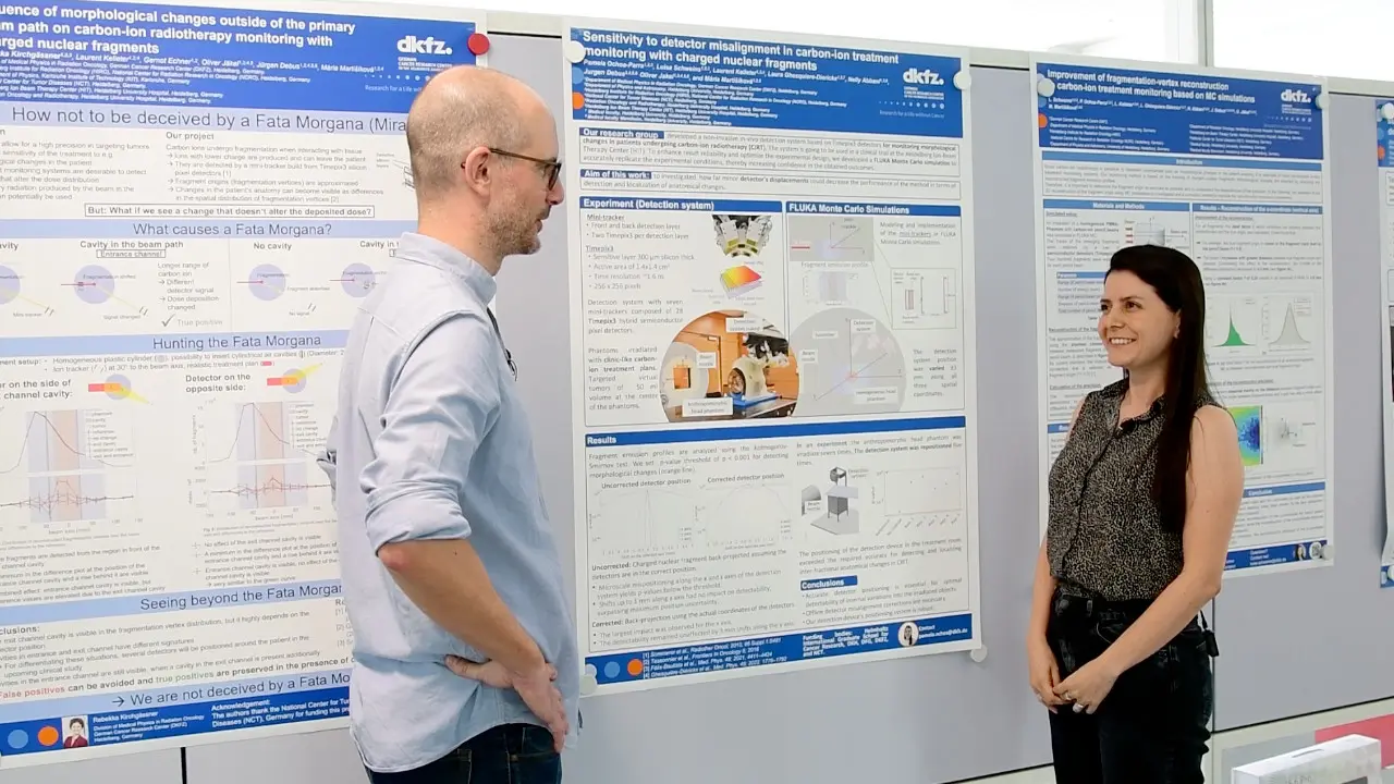 Two researchers engaged in discussion in front of research posters. One person, wearing glasses and a light sweater, stands on the left, while the other person, in a sleeveless top, smiles and listens. The posters contain scientific information related to their research.