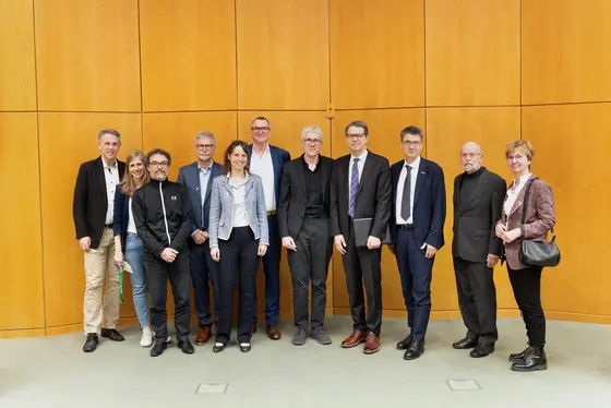 Eine Gruppe von zwölf Personen steht in einem Raum mit Holzverkleidung. Sie sind akademisch gekleidet und wirken feierlich. Im Hintergrund sind keine weiteren Details erkennbar. Die Personen scheinen an einer Veranstaltung zu einem Preis beteiligt zu sein, möglicherweise im wissenschaftlichen Bereich.