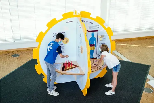 Zwei Kinder spielen an gelber Mitmachstation mit Infotafeln und Lernspielen zu Viren und HPV.