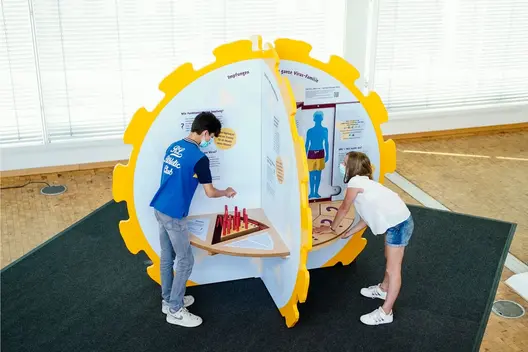 Zwei Kinder spielen an gelber Mitmachstation mit Infotafeln und Lernspielen zu Viren und HPV.