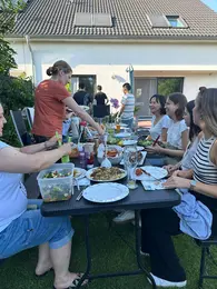 Eine Gruppe von Menschen sitzt an einem Tisch im Freien und genießt ein gemeinsames Essen. Eine Frau serviert verschiedene Speisen, während andere freundlich miteinander lachen und sich unterhalten. Der Tisch ist reich gedeckt mit Essen und Getränken.
