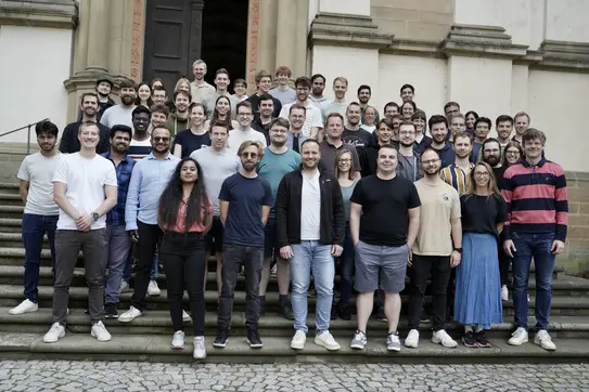 Eine große Gruppe von Menschen steht auf einer Treppe vor einem Gebäude. Sie lächeln und posieren gemeinsam für das Foto. Die Gruppe besteht aus Männern und Frauen unterschiedlichen Alters und Herkunft, die in lässiger Kleidung gekleidet sind.
