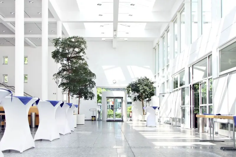 The image shows a spacious, well-lit lobby featuring several white tables draped in blue fabric. Potted trees are positioned along the sides, and large glass doors lead outside. Bright sunlight streams through the windows, enhancing the modern, open atmosphere of the space.