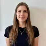 Eine Frau mit langen, glatten, braunen Haaren steht vor einer hellen Wand. Sie trägt ein schwarzes T-Shirt und eine goldene Kette mit einem Anhänger in Form des Buchstabens "D". Ihr Gesicht ist freundlich und ausdrucksvoll.