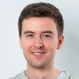 Ein junger Mann lächelt freundlich in die Kamera. Er hat kurzes, dunkelbraunes Haar und trägt ein helles, lässiges Hemd über einem weißen T-Shirt. Der Hintergrund ist hell und unauffällig.