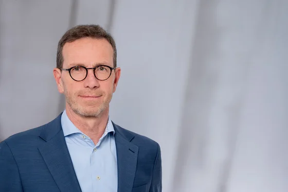 Ein Mann mit Brille und kurzem Haar trägt einen blauen Anzug und ein hellblaues Hemd. Er steht vor einem verschwommenen, hellen Hintergrund und blickt neutral in die Kamera.