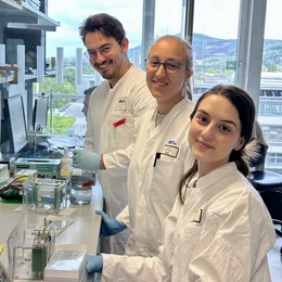 Drei Personen in Laborkleidung stehen an einem Tisch in einem Labor. Sie lächeln und arbeiten mit wissenschaftlichen Instrumenten. 