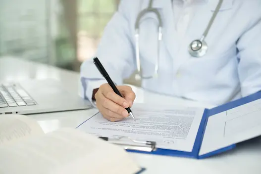 A doctor in a white coat is seated at a desk, writing on a document with a pen. A stethoscope is draped around their neck. A laptop and an open book are nearby, while a clipboard holds additional paperwork.