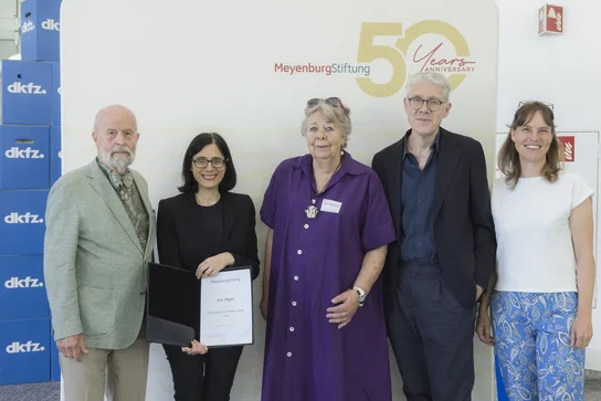 Fünf Personen stehen lächelnd vor einem Banner, das das 50-jährige Jubiläum der Meyenburg Stiftung feiert. Eine Frau in der Mitte hält ein Dokument in der Hand. Die anderen Personen sind in formeller Kleidung gekleidet. Im Hintergrund sind einige Kartons sichtbar.