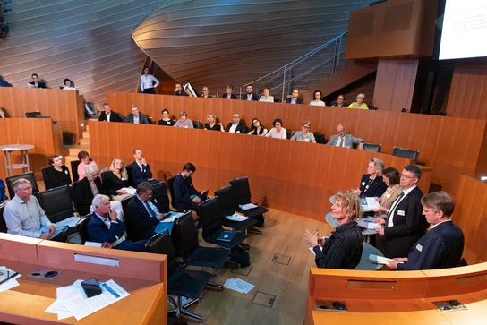 Eine Versammlung oder Konferenz in einem modernen Saal. Im Vordergrund diskutieren mehrere Personen, während im Hintergrund ein Publikum sitzt, das aufmerksam zuhört. Die Umgebung ist hell erleuchtet und hat eine holzverkleidete Architektur.