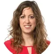 A woman with wavy brown hair smiles warmly. She is wearing a bright red top, with a circular crop around her face, which highlights her friendly expression. The background is plain, drawing focus to her face.
