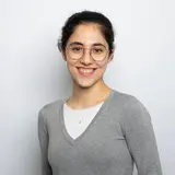 Eine junge Frau mit braunen Haaren und einer Brille lächelt in die Kamera. Sie trägt ein graues Oberteil über einem weißen Shirt und steht vor einem hellen Hintergrund.