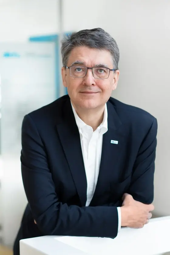 Ein Mann mittleren Alters mit kurzen grauen Haaren steht vor einem hellen Hintergrund. Er trägt einen dunklen Anzug und eine weiße Hemdbluse. Mit einer Hand stützt er seinen Ellenbogen auf eine weiße Fläche, während er in die Kamera lächelt. Die Atmosphäre ist professionell und einladend.