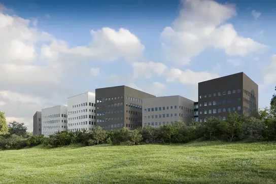Mehrteiliges, modernes Gebäudekomplex mit rechteckigen Fassaden in unterschiedlichen Grautönen. Im Vordergrund eine grüne Wiese mit Büschen.