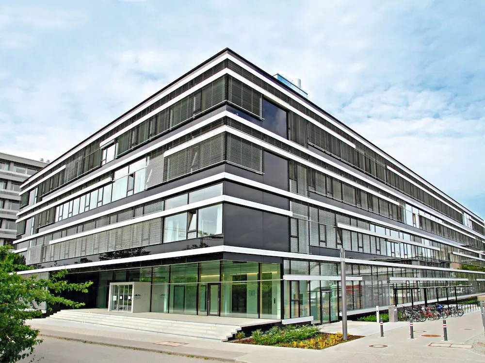 Ein modernes, mehrstöckiges Gebäude mit vielen Glasfenstern und schwarzen Fassadenelementen. Die Eingangszone ist großzügig gestaltet, mit Treppenstufen und einem gläsernen Eingang. Im Vordergrund sind einige Fahrradständer sichtbar. Der Himmel ist teilweise bewölkt.