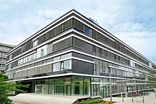 Ein modernes, mehrstöckiges Gebäude mit vielen Glasfenstern und schwarzen Fassadenelementen. Die Eingangszone ist großzügig gestaltet, mit Treppenstufen und einem gläsernen Eingang. Im Vordergrund sind einige Fahrradständer sichtbar. Der Himmel ist teilweise bewölkt.