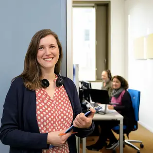 Eine lächelnde Frau mit langen, braunen Haaren steht im Vordergrund und hält einen Stift und Unterlagen. Im Hintergrund sind zwei weitere Personen an Computerarbeitsplätzen zu sehen, die Kopfhörer tragen und miteinander kommunizieren. Die Umgebung wirkt modern und freundlich.