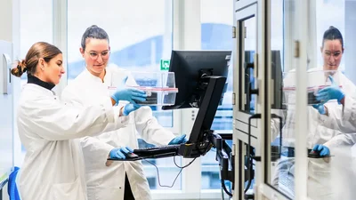 Zwei Wissenschaftlerinnen in Laborkitteln arbeiten zusammen an einem Computer. Eine hält einen durchsichtigen Behälter mit einem roten Deckel, während die andere auf den Bildschirm zeigt. Im Hintergrund sind weitere Laborgeräte sichtbar.