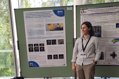 Eine Person steht neben zwei wissenschaftlichen Posterpräsentationen in einem hellen Raum mit Fenster. Die Person trägt eine blaue Bluse und beige Hose und hält einen Notizblock in der Hand. Im Hintergrund sind Grafiken und Informationen auf den Postern zu sehen.