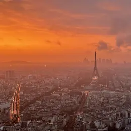 A panoramic view of Paris at sunset, showcasing the Eiffel Tower silhouetted against an orange and cloudy sky. The city's streets reflect the fading light, creating a tranquil yet vibrant atmosphere. The scene captures the essence of Paris as day transitions to night.