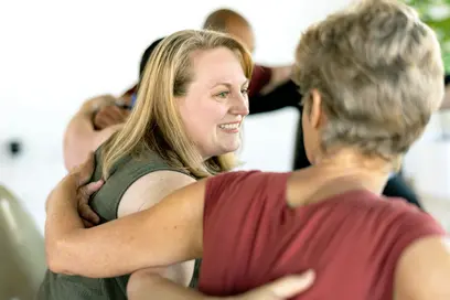 Eine Frau lächelt und umarmt eine andere Frau in einer freundlichen, entspannten Atmosphäre. Sie scheinen an einer Gruppenaktivität teilzunehmen, die positive zwischenmenschliche Interaktionen fördert. Die Gesichter sind freundlich und offen, und die Verbindung zwischen den Personen ist spürbar.
