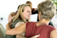 Eine Frau lächelt und umarmt eine andere Frau in einer freundlichen, entspannten Atmosphäre. Sie scheinen an einer Gruppenaktivität teilzunehmen, die positive zwischenmenschliche Interaktionen fördert. Die Gesichter sind freundlich und offen, und die Verbindung zwischen den Personen ist spürbar.