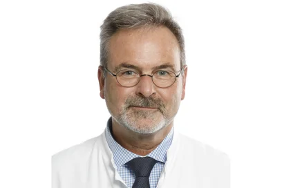 Ein Mann mit Brille und grauem Haar, der einen weißen Laborkittel trägt. Er hat einen freundlichen Gesichtsausdruck und ist vor einem neutralen Hintergrund abgebildet.