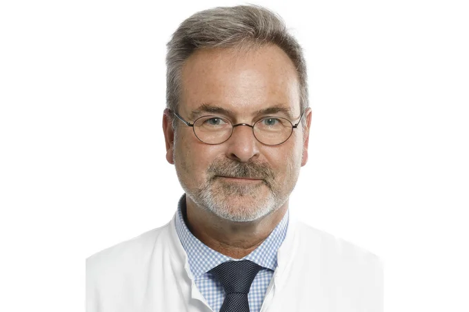 Ein Mann mit Brille und grauem Haar, der einen weißen Laborkittel trägt. Er hat einen freundlichen Gesichtsausdruck und ist vor einem neutralen Hintergrund abgebildet.