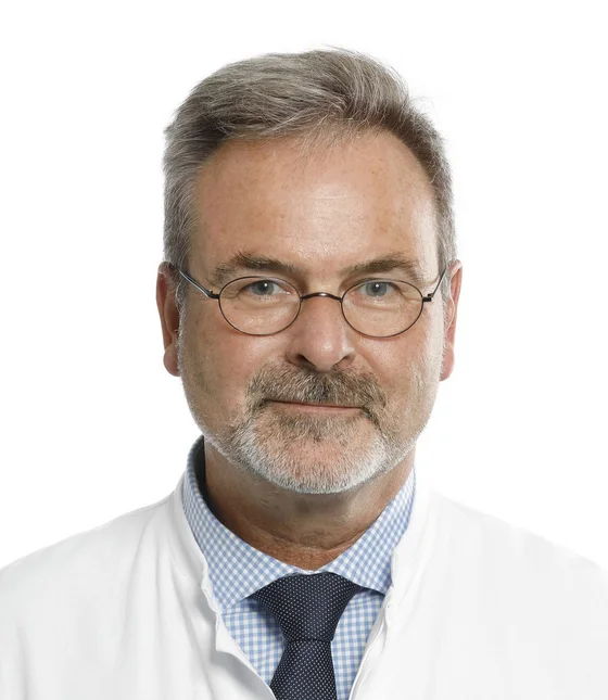 Ein Mann mit Brille und grauem Haar, der einen weißen Laborkittel trägt. Er hat einen freundlichen Gesichtsausdruck und ist vor einem neutralen Hintergrund abgebildet.