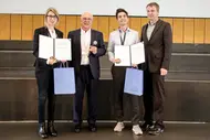 Vier Personen stehen vor einer Bühne. Zwei halten Urkunden in den Händen. Alle vier Personen lächeln. Die Szene vermittelt eine feierliche Atmosphäre, im Rahmen einer Auszeichnung oder Zeremonie.