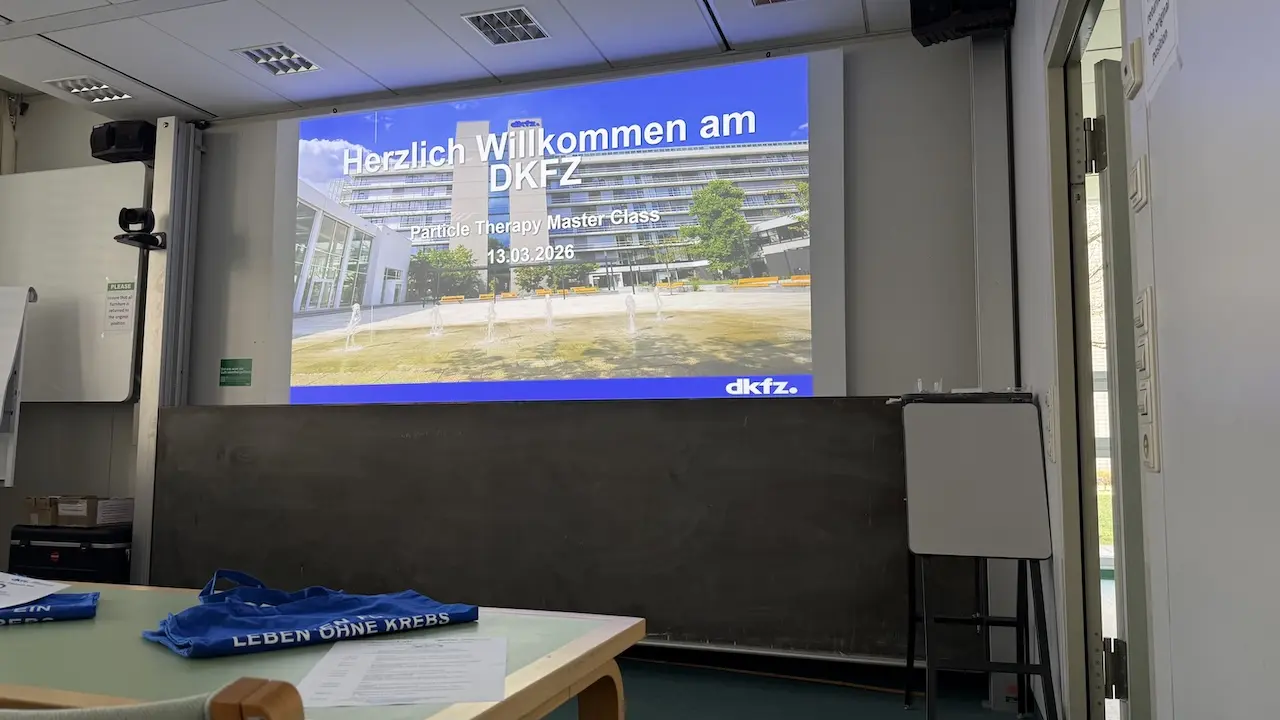 Ein moderner Seminaraum mit einer Präsentation an der Wand, die "Herzlich Willkommen am DKFZ" zeigt. Auf einem Tisch liegen blaue Taschen, und eine Tafel steht im Hintergrund. Das Bild vermittelt eine einladende Atmosphäre für eine Veranstaltung.