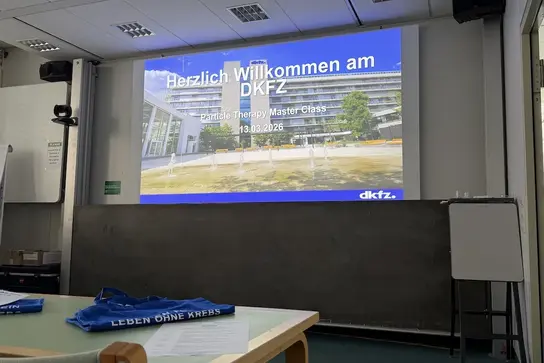 Ein moderner Seminaraum mit einer Präsentation an der Wand, die "Herzlich Willkommen am DKFZ" zeigt. Auf einem Tisch liegen blaue Taschen, und eine Tafel steht im Hintergrund. Das Bild vermittelt eine einladende Atmosphäre für eine Veranstaltung.