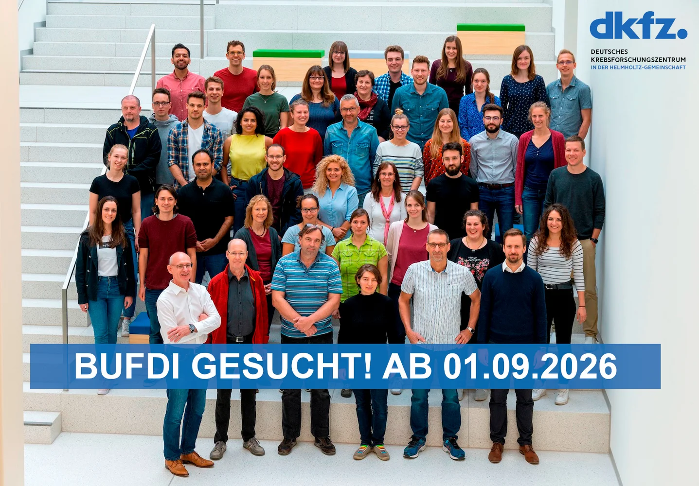 Gruppenfoto von 43 Personen auf einer Treppe, die vor einem hellen Hintergrund stehen. Einige Personen tragen unterschiedliche Kleidung. Im Vordergrund steht ein großer, blauer Text: "BUFDI GESUCHT! AB 01.09.2026".