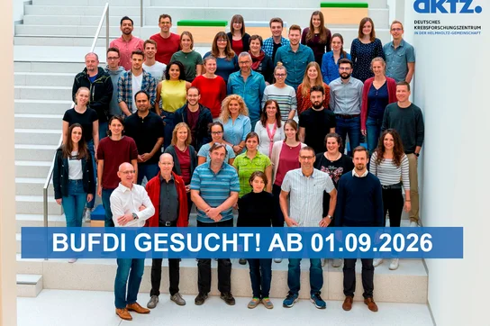 Gruppenfoto von 43 Personen auf einer Treppe, die vor einem hellen Hintergrund stehen. Einige Personen tragen unterschiedliche Kleidung. Im Vordergrund steht ein großer, blauer Text: "BUFDI GESUCHT! AB 01.09.2026".