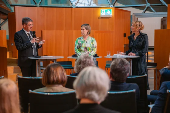 Drei Personen stehen an kleinen Tischen in einem modernen Veranstaltungsraum. Ein Mann spricht, während zwei Frauen ihm zuhören. Im Vordergrund sind unscharfe Silhouetten von Zuhörern zu sehen.