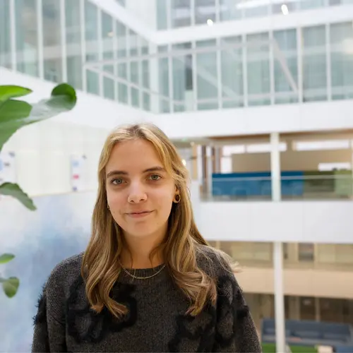 Eine Person mit langen, blonden Haaren steht in einem modern gestalteten Innenraum. Im Hintergrund sind große Fenster und eine Pflanze zu sehen, die dem Raum eine freundliche Atmosphäre verleiht. Die Person trägt einen dunklen Pullover und schaut in die Kamera.