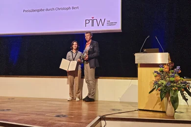 Eine Frau und ein Mann stehen auf einer Bühne. Die Frau hält einen Preis und eine Urkunde in den Händen. Der Mann überreicht den Preis. Im Hintergrund ist ein Podium mit einem blumengeschmückten Tisch und einem Bildschirm sichtbar, auf dem "PTW-Posterpreis" angezeigt wird.