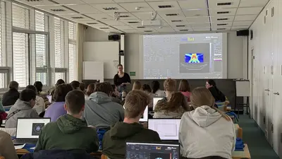 Eine Lehrerin steht vor einer Präsentation in einem Klassenraum, während Schüler an Computern sitzen und auf den Bildschirm schauen. Die Schüler sind von hinten sichtbar und verfolgen die Unterrichtsinhalte.