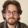 Ein junger Mann mit lockigem, dunklem Haar und einer Brille lächelt in die Kamera. Er trägt einen grauen Pullover mit einem besonderen Kragen. Der Hintergrund ist hell und schlicht.