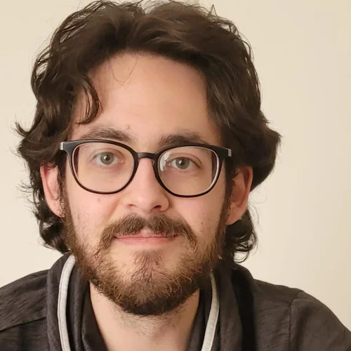 Ein junger Mann mit lockigem, dunklem Haar und einer Brille lächelt in die Kamera. Er trägt einen grauen Pullover mit einem besonderen Kragen. Der Hintergrund ist hell und schlicht.