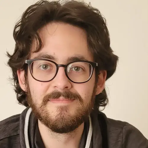 Ein junger Mann mit lockigem, dunklem Haar und einer Brille lächelt in die Kamera. Er trägt einen grauen Pullover mit einem besonderen Kragen. Der Hintergrund ist hell und schlicht.